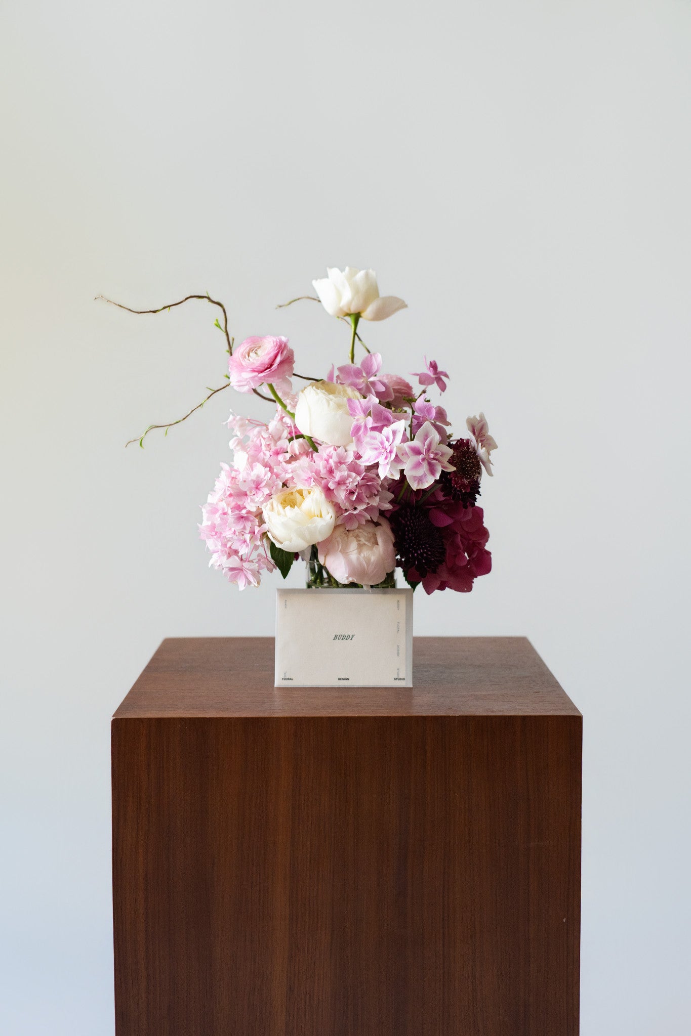 A floral arrangement in a white rectangular box, placed on a dark brown wooden pedestal against a light gray background.