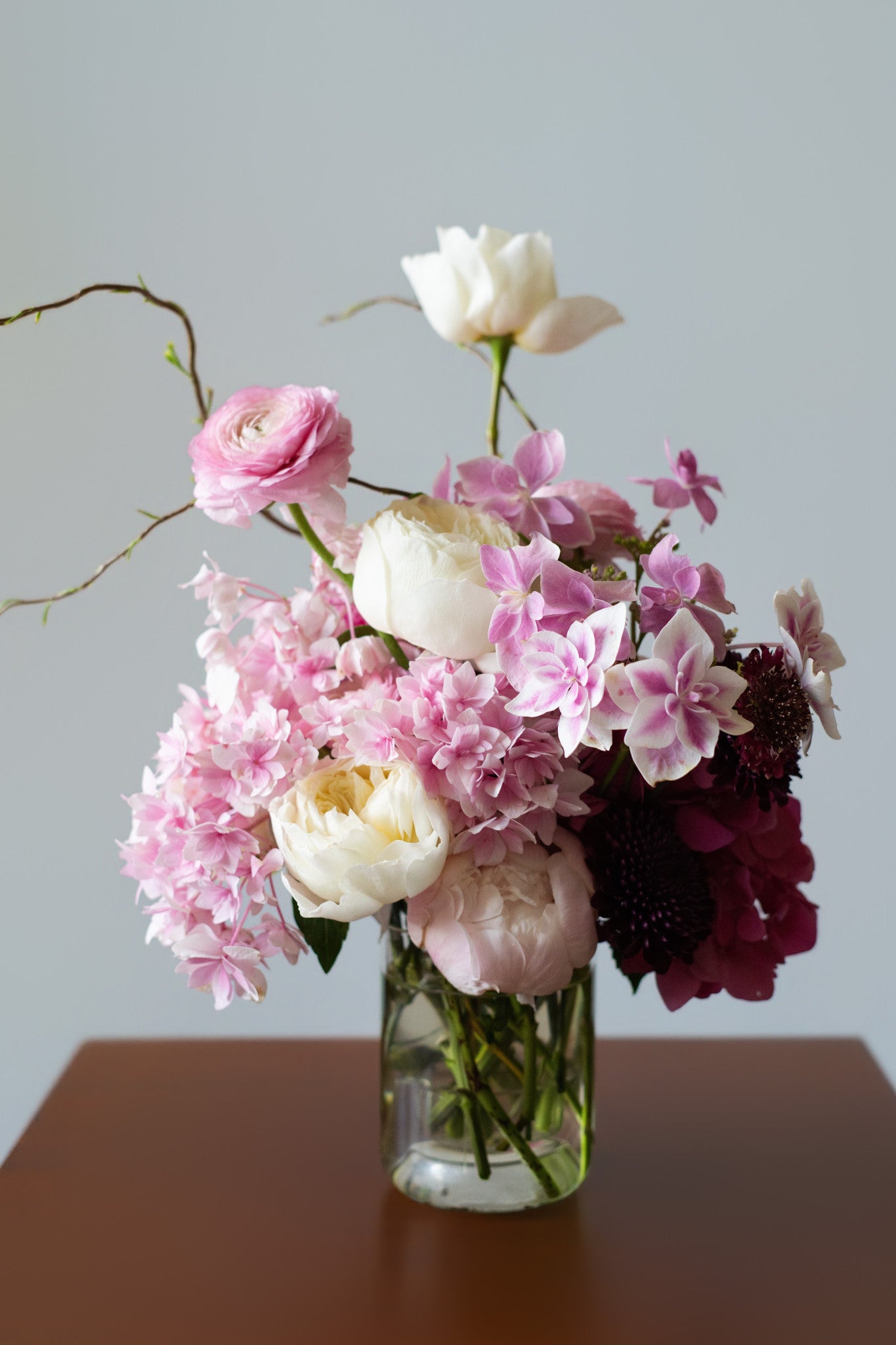 A mixed floral arrangement in a clear glass vase, featuring pink and white flowers with dark burgundy accents, set on a brown wooden surface against a light gray background.