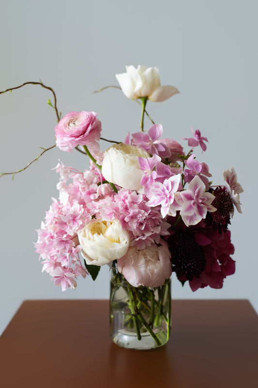 A mixed floral arrangement in a clear glass vase, featuring pink and white flowers with dark burgundy accents, set on a brown wooden surface against a light gray background.