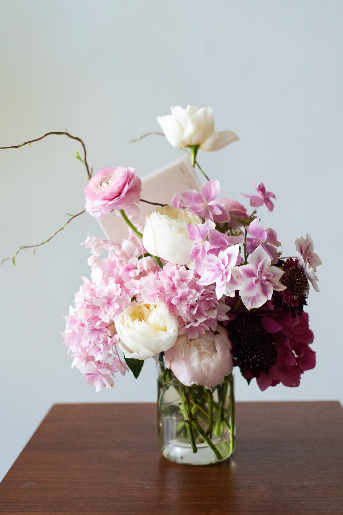 A floral arrangement of pink, white, and deep burgundy flowers in a clear glass vase, placed on a dark wooden table against a light gray wall.