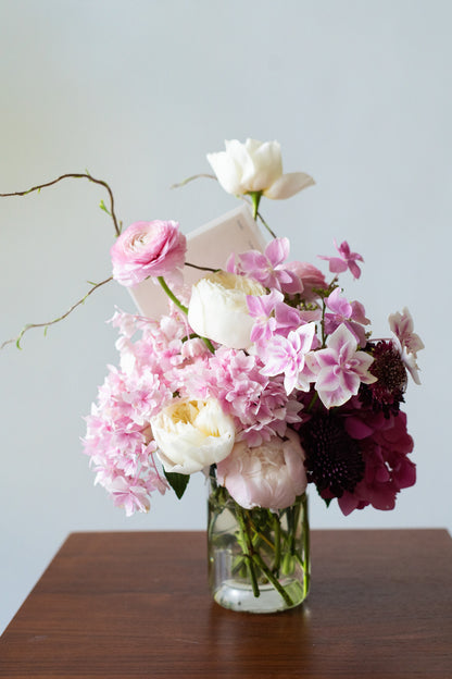 A floral arrangement of pink, white, and deep burgundy flowers in a clear glass vase, placed on a dark wooden table against a light gray wall.