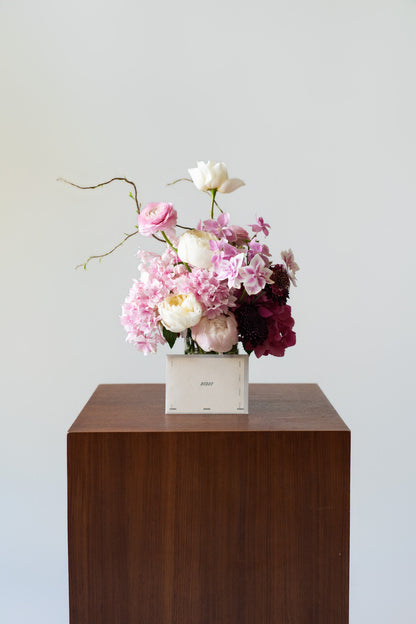 A floral arrangement in a white rectangular box, placed on a dark brown wooden pedestal against a light gray background.
