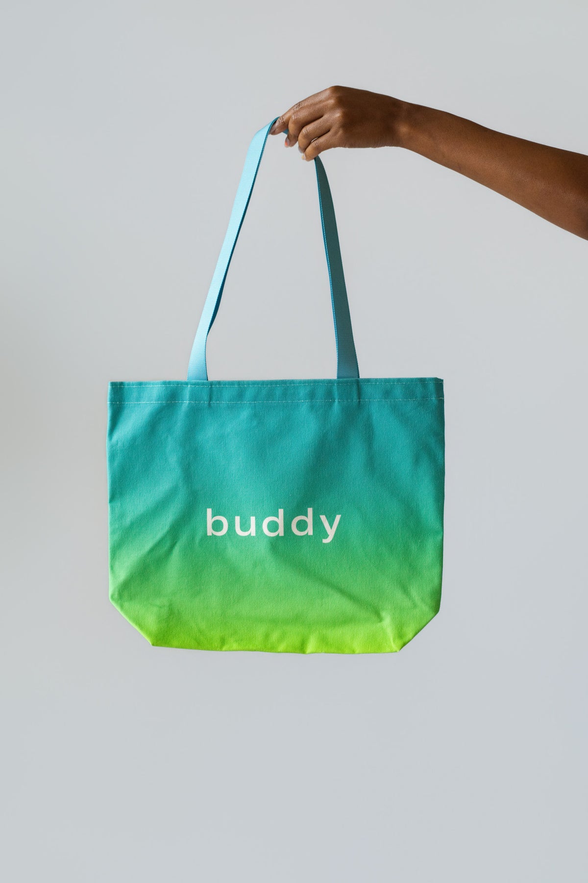A hand holding a canvas tote bag with a teal-to-yellow gradient and the word 'buddy' printed in white on the front, set against a solid light gray background.