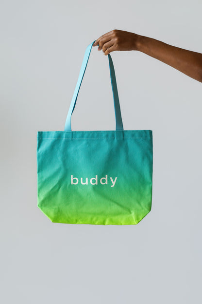 A hand holding a canvas tote bag with a teal-to-yellow gradient and the word 'buddy' printed in white on the front, set against a solid light gray background.