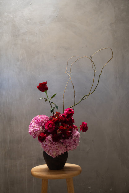 A floral arrangement in a dark vase placed on a small wooden stool against a textured gray wall. The bouquet includes pink hydrangea-like blooms, deep red roses, and thin curly twigs extending upward.
