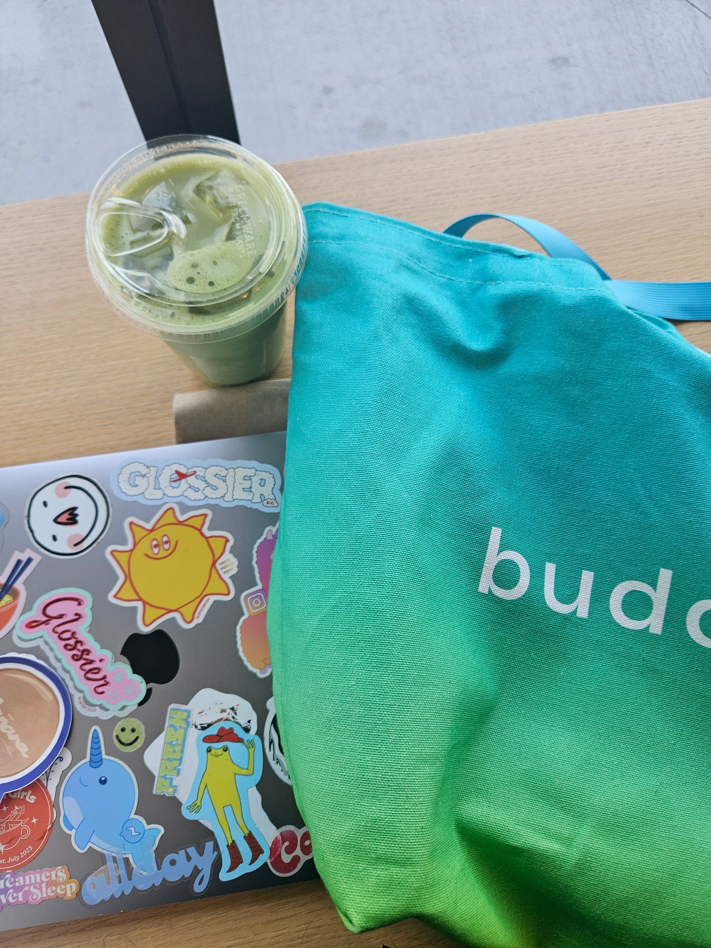 Green smoothie in a cup, laptop with stickers, and ombre turquoise bag on a wooden surface.