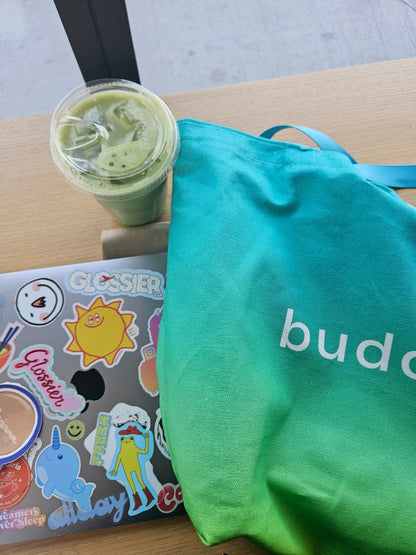 Green smoothie in a cup, laptop with stickers, and ombre turquoise bag on a wooden surface.
