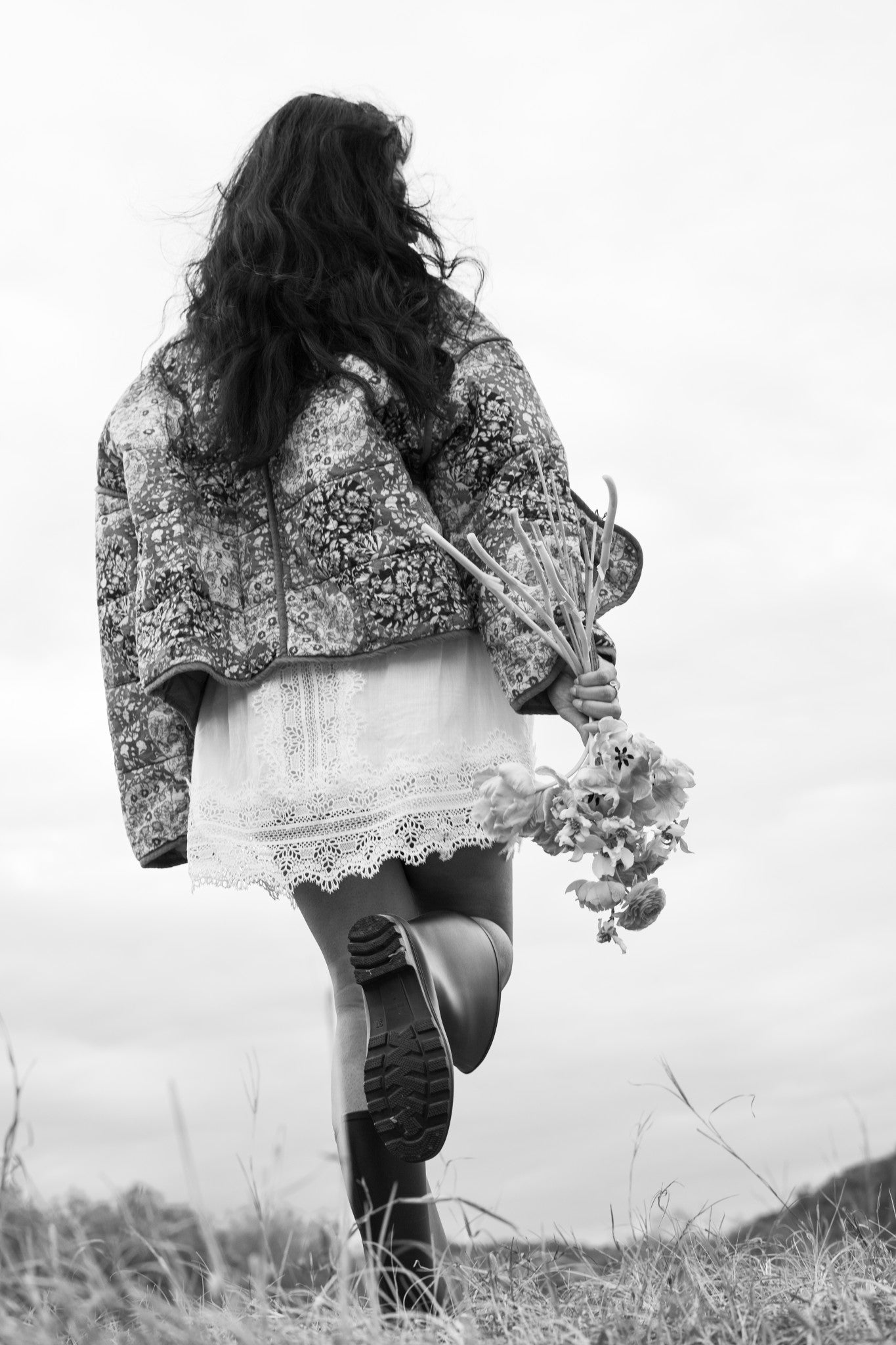 Person wearing a patterned coat and skirt with a bouquet of flowers, standing on grass with a blurred background.