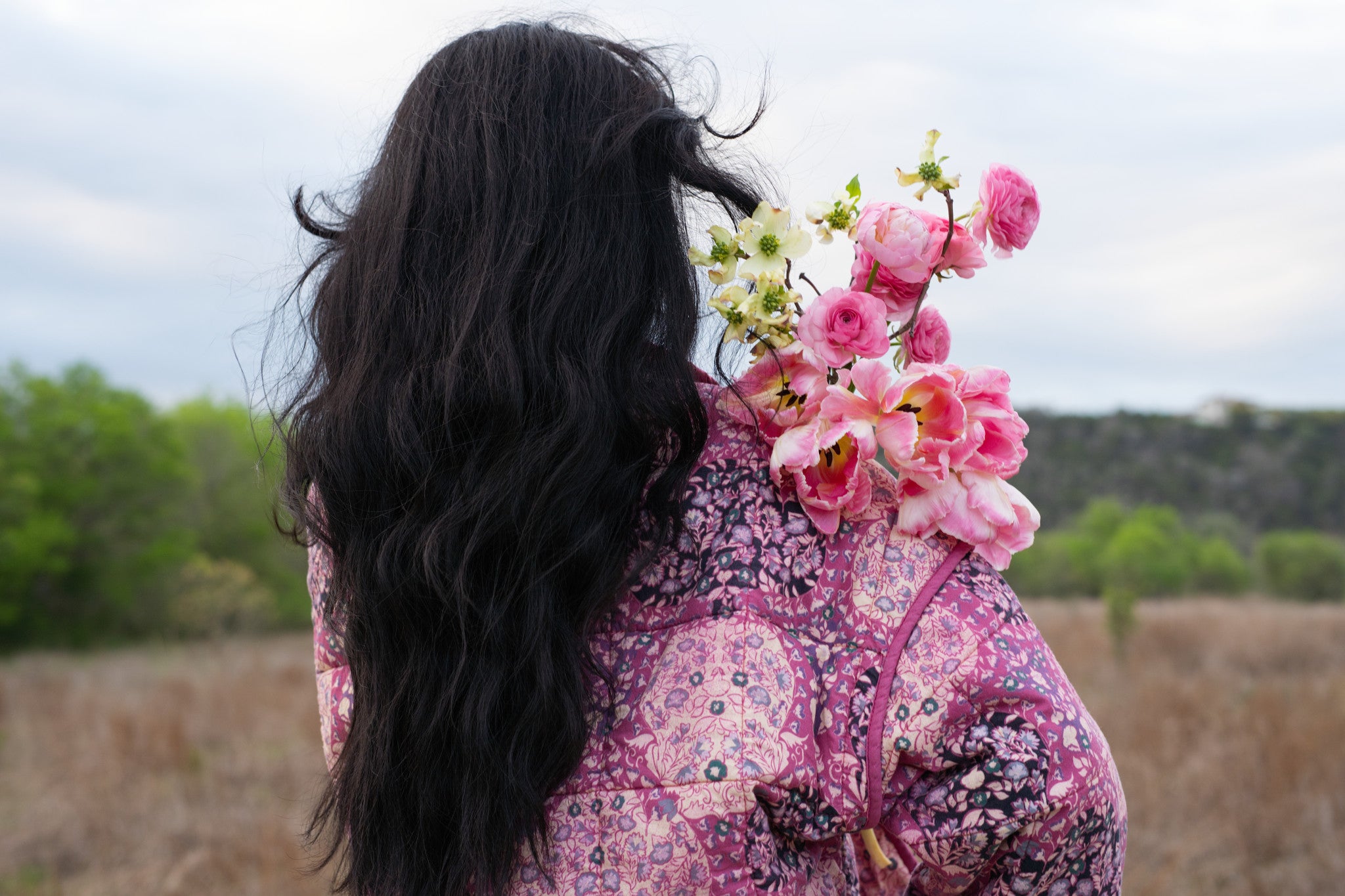 Person with long dark hair and a pink floral jacket standing in an open field in Austin Texas.